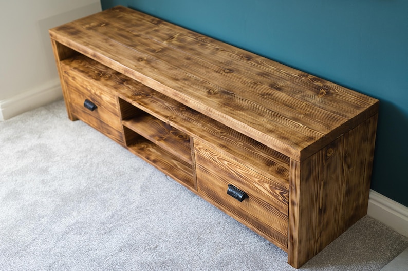 May include: A wooden TV stand with a warm brown finish. The stand features two drawers with black handles and open shelving. The wood grain is visible, adding texture. The stand is placed on a light gray carpet, with a teal wall in the background.