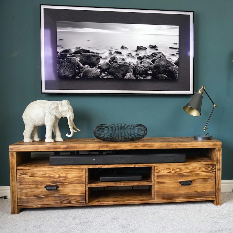 May include: A rustic wooden TV stand with two drawers and open shelving. The stand is topped with a white elephant figurine, a dark blue decorative bowl, and a black lamp. A framed black and white landscape photo hangs above.