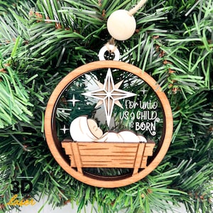 May include: Circular wooden Christmas ornament with a star and the text "For unto us a child is born". The ornament depicts a baby in a manger. The ornament is hanging on a Christmas tree. The frame is approximately 10 cm in diameter.