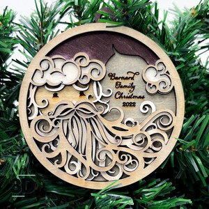 May include: A round wooden Christmas ornament with a laser-cut design of Santa Claus's face. The ornament is decorated with swirls and the text "Barnard Family Christmas 2022".