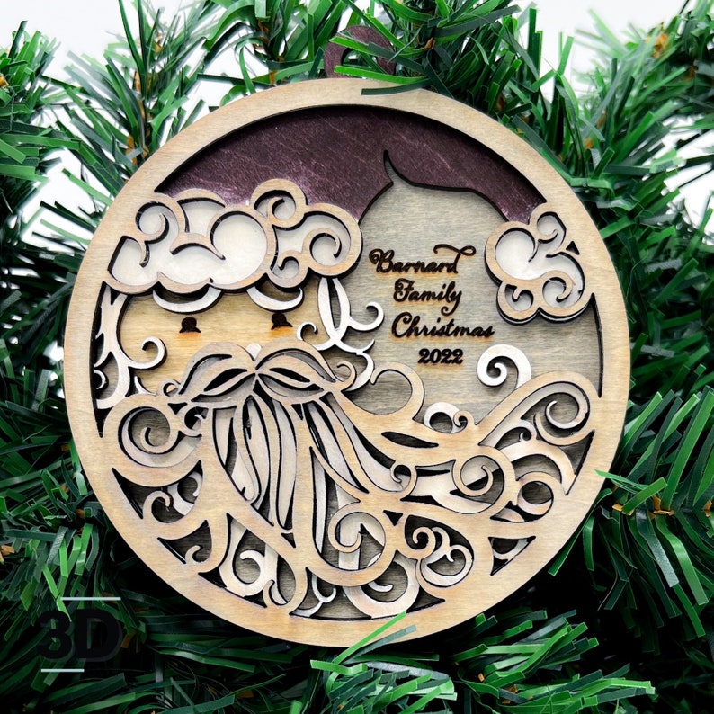 May include: A round wooden Christmas ornament with a laser-cut design of Santa Claus's face. The ornament is decorated with swirls and the text "Barnard Family Christmas 2022".