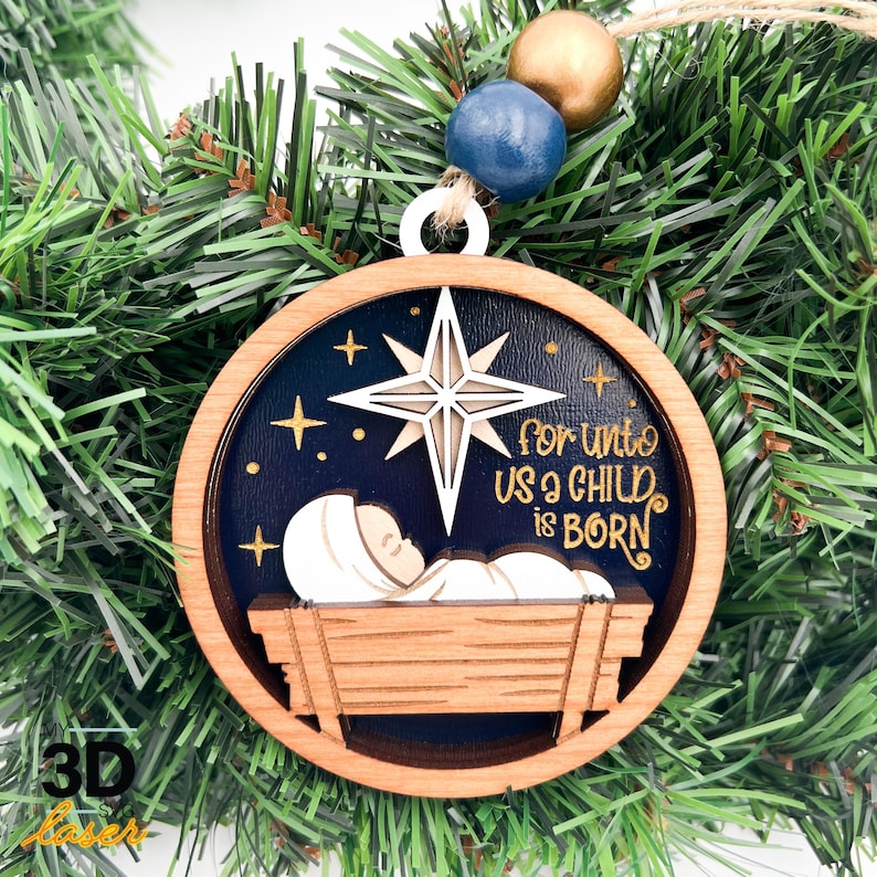 May include: Wooden Christmas bauble with a layered design. The bauble features a star, the text "For Unto Us a Child is Born", and a depiction of baby Jesus in a manger. The bauble is hanging on a Christmas tree.