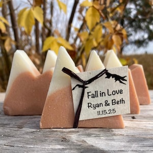 May include: Mountain-shaped soaps in shades of cream and peach, arranged in a group. A small tag tied with a dark ribbon reads "Fall in Love Ryan & Beth 11.15.25". The soaps are on a wooden surface, with blurred foliage in the background.