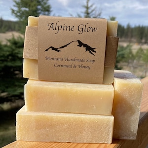 May include: Four bars of handmade soap stacked on top of each other. The top bar has a brown paper label that reads "Alpine Glow Montana Handmade Soap Cornmeal & Honey".