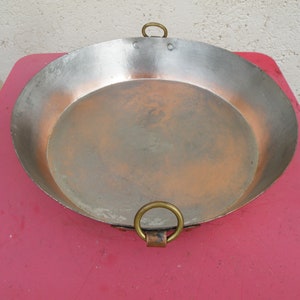 May include: A large, round copper pan with two brass handles. The pan is slightly tarnished and has a worn appearance.