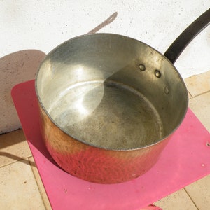 May include: A copper pot with a black handle. The pot is sitting on a pink surface. The pot has a hammered finish and is empty.