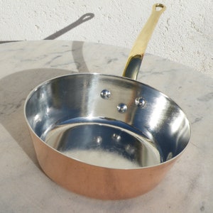 May include: A copper saucepan with a brass handle. The saucepan has a shiny, silver interior and three rivets on the side.