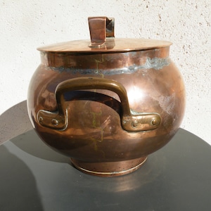 Puede incluir: Una olla grande de cobre con tapa y una sola asa. La olla tiene un cuerpo redondeado y una base ancha. El asa es de latón y tiene un diseño decorativo. La olla está sobre una superficie oscura.