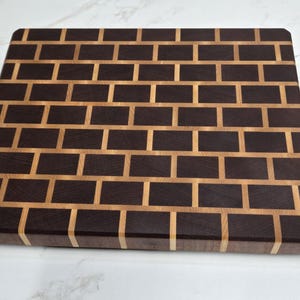May include: A rectangular wooden cutting board with a brick pattern made from dark brown and light brown wood.