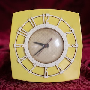 May include: A vintage yellow square clock with a white circular face and white Roman numerals. The clock hands are black and red. The clock has a retro design.