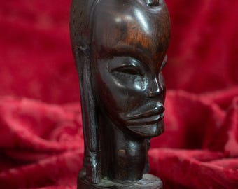 May include: A dark brown wooden sculpture of a stylized human head. The sculpture has a smooth, polished finish with carved facial features, including closed eyes and a defined mouth. The head sits atop a cylindrical base.