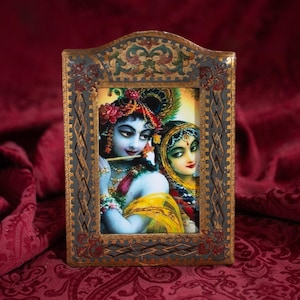 May include: A framed artwork depicting two figures with blue and yellow skin tones, adorned with floral headpieces and jewelry. The frame is ornate, with a dark, textured finish and gold accents. The background is a deep red, patterned fabric.