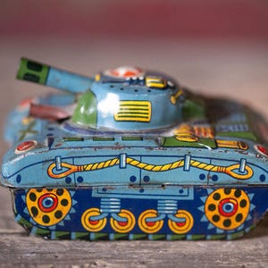 May include: A vintage-style tin toy tank featuring a light blue body with green accents, a rotating turret, and a long barrel. The sides are decorated with yellow rope illustrations and yellow wheels with red centers on a dark blue background.