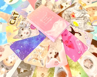 May include: A set of colorful tarot cards featuring illustrations of cats and kittens. The cards are laid out in a fan shape, with the box for the deck in the center. The box is pink and has the words "Kittens and Kittie Lenormand" printed on it.