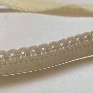 May include: A close-up of a cream-colored elastic trim with a delicate scalloped edge. The trim is soft and flexible, making it ideal for adding a touch of elegance to sewing projects.
