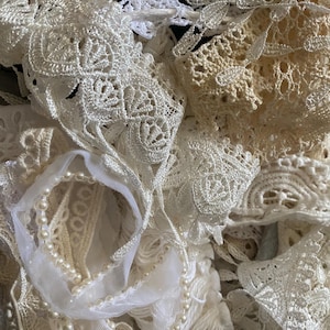 May include: A close-up of various white and cream-colored lace and ribbons. The image features different textures and patterns of lace, including floral and geometric designs. A strand of faux pearls is also visible, adding a touch of elegance to the composition.