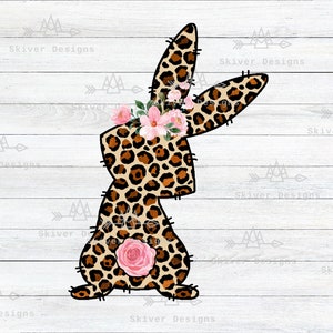 May include: A brown and tan leopard print bunny with a pink flower crown and a pink rose on its back. The bunny is sitting on a white wood background.