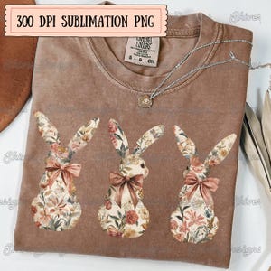 May include: A brown t-shirt with three floral bunny silhouettes. The bunnies are in a row, facing the viewer. The bunnies are made of a floral pattern with pink, green, and yellow flowers. The t-shirt is a sublimation print, meaning the design is printed directly onto the fabric.