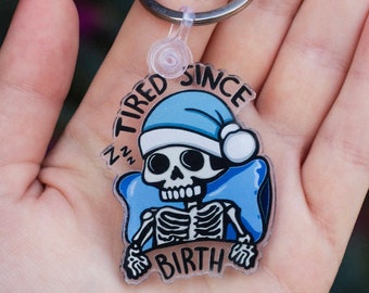 May include: A clear acrylic keychain featuring a cartoon skeleton wearing a blue Santa hat and sleeping on a blue pillow. The text "TIRED SINCE BIRTH" is printed on the keychain.