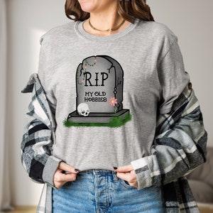 May include: A grey long-sleeved shirt with a graphic of a tombstone that reads "RIP My Old Hobbies".