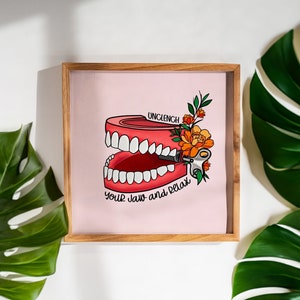May include: A framed print with a pink background featuring a cartoon illustration of a set of dentures with a key winding up the jaw. The text "Unclench your jaw and relax" is below the dentures. The print is surrounded by a wooden frame.