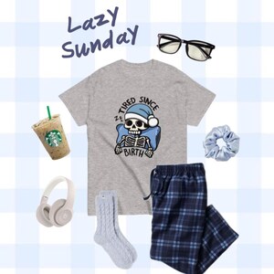 May include: A gray t-shirt with a skeleton wearing a Santa hat and the text "Tired Since Birth". The outfit also includes blue and white plaid pajama pants, a pair of gray socks, a light blue hair scrunchie, a pair of black glasses, and a white cup of coffee.