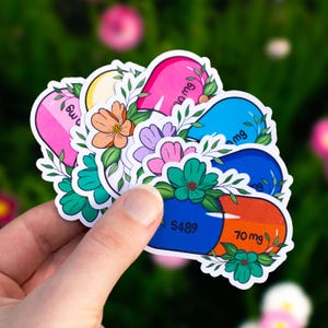 May include: A set of five colorful pill-shaped stickers with floral designs. Each sticker features a different color pill with a dosage amount printed on it, such as "70 mg" and "60 mg".