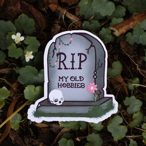 May include: A gray tombstone with a white skull and pink flower. The tombstone reads "RIP My Old Hobbies".