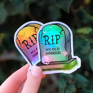 Può includere: Due adesivi a forma di lapide. Uno ha il testo "RIP My Old Hobbies" e l'altro "RIP My Old Hopes". Presentano un gradiente arcobaleno e un piccolo teschio. Gli adesivi sono tenuti in mano.