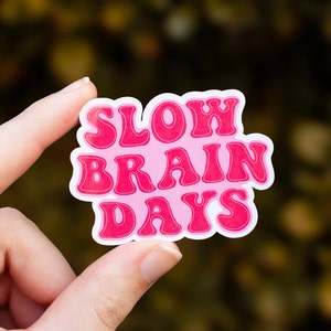 May include: Pink sticker with the text "SLOW BRAIN DAYS" in a retro font style.