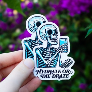 Könnte beinhalten: Ein Aufkleber mit zwei blauen Skeletten, die Wasser aus einem Glas trinken. Der Text auf dem Aufkleber lautet "Hydrate or Die-Drate".