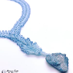 Könnte beinhalten: Eine zarte blaue Perlenkette mit einem hellblauen Kristallanhänger. Die Kette besteht aus kleinen, abgestuften Perlen, die einen kaskadierenden Effekt erzeugen. Der Kristallanhänger ist unregelmäßig geformt und hat eine glitzernde, irisierende Oberfläche.