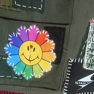May include: A green canvas jacket with a rainbow flower patch with a smiley face and a red blood splatter. The jacket has other patches including a black patch with white stitching and a black patch with a white logo that says "STAR TREK". The jacket also has a white patch with black text that says "TIM HENDRIX KURT COBAIN JANKE TOPLIN TIM".