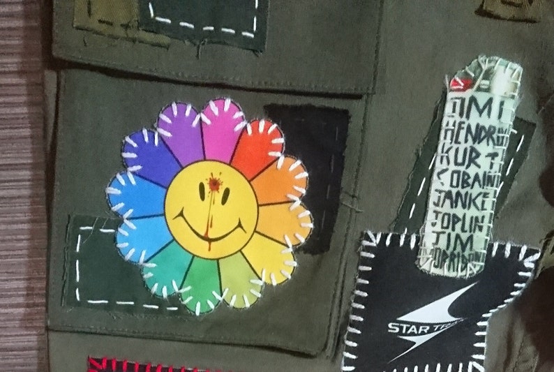 May include: A green canvas jacket with a rainbow flower patch with a smiley face and a red blood splatter. The jacket has other patches including a black patch with white stitching and a black patch with a white logo that says "STAR TREK". The jacket also has a white patch with black text that says "TIM HENDRIX KURT COBAIN JANKE TOPLIN TIM".