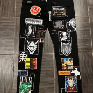 Puede incluir: Vaqueros negros de denim con varios parches y diseños gráficos, incluyendo una calavera, una máscara de demonio y el texto "AKIRA".