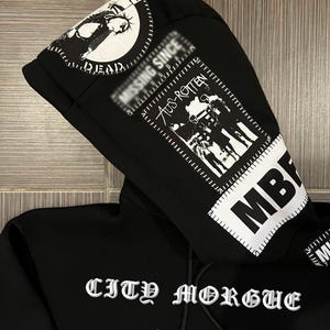 Pu&ograve; includere: Felpa con cappuccio nera con stampa grafica bianca. La stampa include il testo "MISSING SINCE", "DEAD", "AUS-ROTTEN", "MB" e "CITY MORGUE".