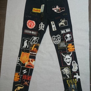 May include: Black denim pants with various patches sewn on, including a skull, a hand holding a gun, a red fish, and a red devil mask. The patches are in different sizes and shapes, and some have text on them, such as "City Morgue" and "Welcome to New York".