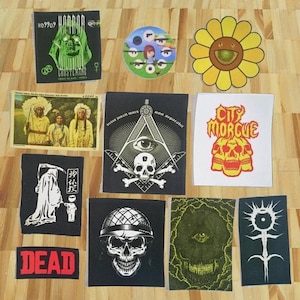 May include: A collection of iron-on patches featuring various designs, including a skull and crossbones, a smiley face, a grim reaper, and a city morgue logo. The patches are on a wooden surface.