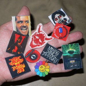 Puede incluir: Una colección de parches para planchar con varios diseños, incluyendo una calavera, una cara sonriente, una píldora, un póster de película y un personaje de una película de terror. Los parches están en una mano, listos para plancharse sobre tela.