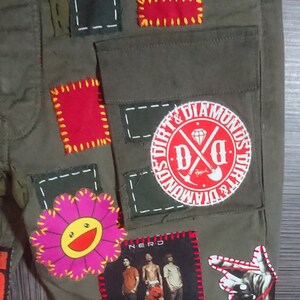 May include: Green cargo pants with various patches sewn on. Patches include a red circle with the text "Dirt & Diamonds" and the letters "D&D", a pink flower with a yellow smiley face, a red square with yellow stitching, a black square with white stitching, a black and white photo of a band with the word "Nerd" above it, and a black hand with red stitching.
