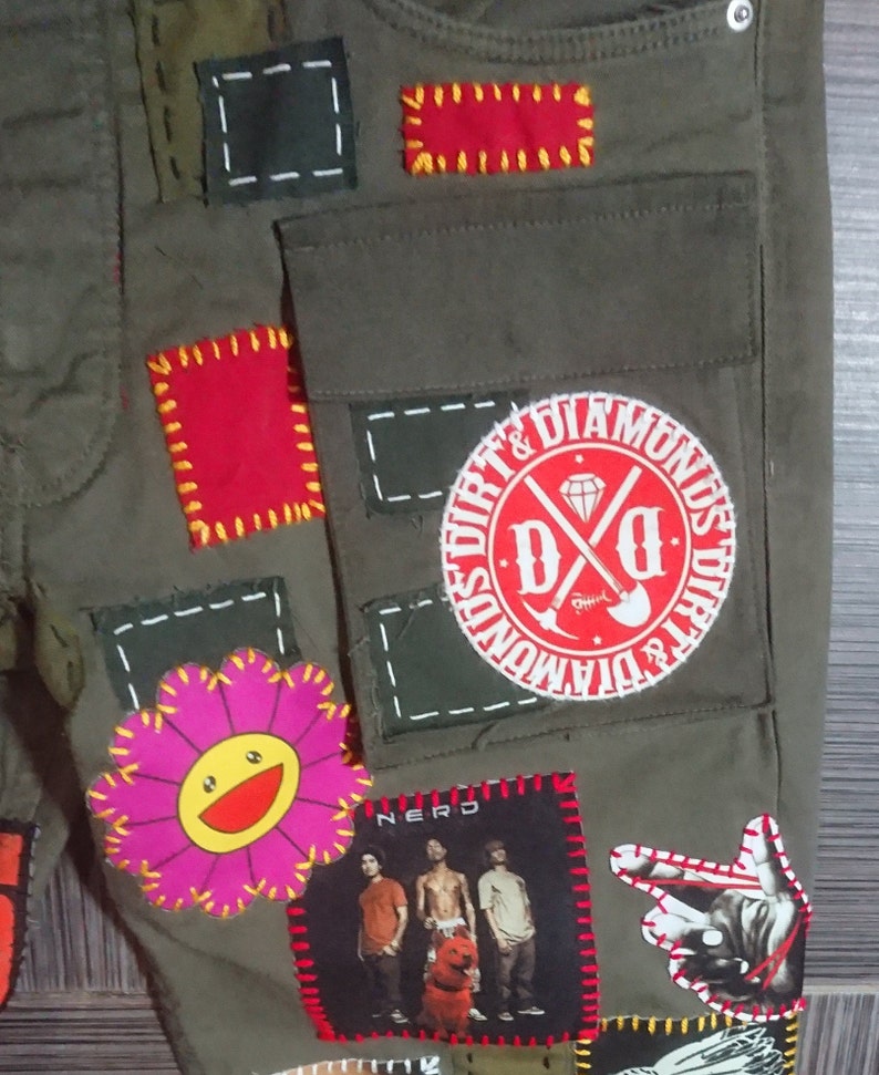 May include: Green cargo pants with various patches sewn on. Patches include a red circle with the text "Dirt & Diamonds" and the letters "D&D", a pink flower with a yellow smiley face, a red square with yellow stitching, a black square with white stitching, a black and white photo of a band with the word "Nerd" above it, and a black hand with red stitching.