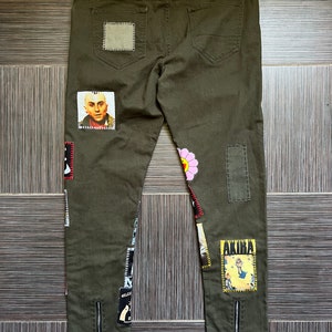 May include: Olive green jeans with various patches, including a flower, a cartoon character, and a portrait of a man. The jeans have zippered hems and a "Sad Sad Sad" patch on the back.