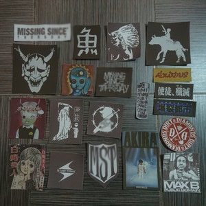 May include: A collection of vintage stickers featuring various designs, including a demon, a skull, a cartoon character, a Native American, a band logo, and a skull with a scythe. The stickers are on a brown wooden surface.