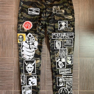 Puede incluir: Pantalones de camuflaje con múltiples parches que muestran varios gráficos y texto. Los parches incluyen una mano sosteniendo una pistola, una cara de diablo y las frases "Intimidate All Witnesses" y "Gone Mad A World".