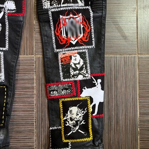 May include: Black denim pants with multiple patches sewn on. The patches feature various designs, including skulls, a cowboy riding a horse, and a person with a red hat. The patches are outlined in white, red, and yellow stitching.