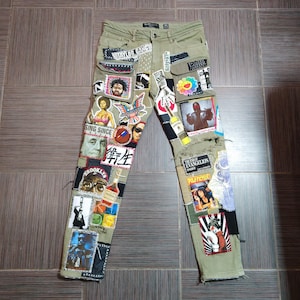 Op de afbeelding: Olijfgroene denim jeans met talrijke patches met portretten, afbeeldingen en tekst. Patches bevatten afbeeldingen van mensen, een bloem en de tekst "Taylor Gang". De jeans hebben een versleten look met scheuren.