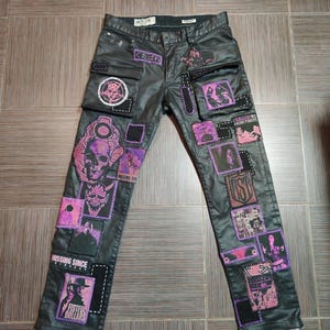 Puede incluir: Pantalones vaqueros negros con múltiples parches en tonos rosas y morados. Los parches presentan varios gráficos, incluyendo calaveras, figuras y texto. Los pantalones tienen un diseño clásico de cinco bolsillos y un lavado oscuro.