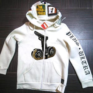 Puede incluir: Sudadera blanca con capucha y cremallera, con un gráfico de pistola negra y plateada en la parte delantera y "VITUS ULTRA" impreso en negro en la manga derecha.