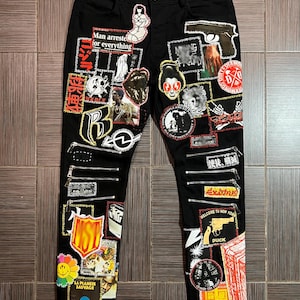 May include: Black denim jeans with various colorful patches and embroidered details. The patches include images, text, and symbols. The text on the patches includes "Man arrested for everything", "Welcome to New York", "Duck", and "La Planete Sauvage".