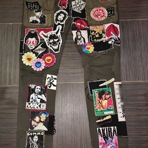 Puede incluir: Pantalón cargo verde con varios parches cosidos, incluyendo imágenes de personas, logotipos y personajes de dibujos animados. Los parches incluyen texto como "DI-HADE", "STARTRAK", "MAXB", "PUBLIC DOMAIN", "GUMMO", "家", "RUFF RYDERS", "DIRE DIARD", "DIRTA DIALD", "マ・ワ振h Masters", "Newport", "DAY", "NCE".